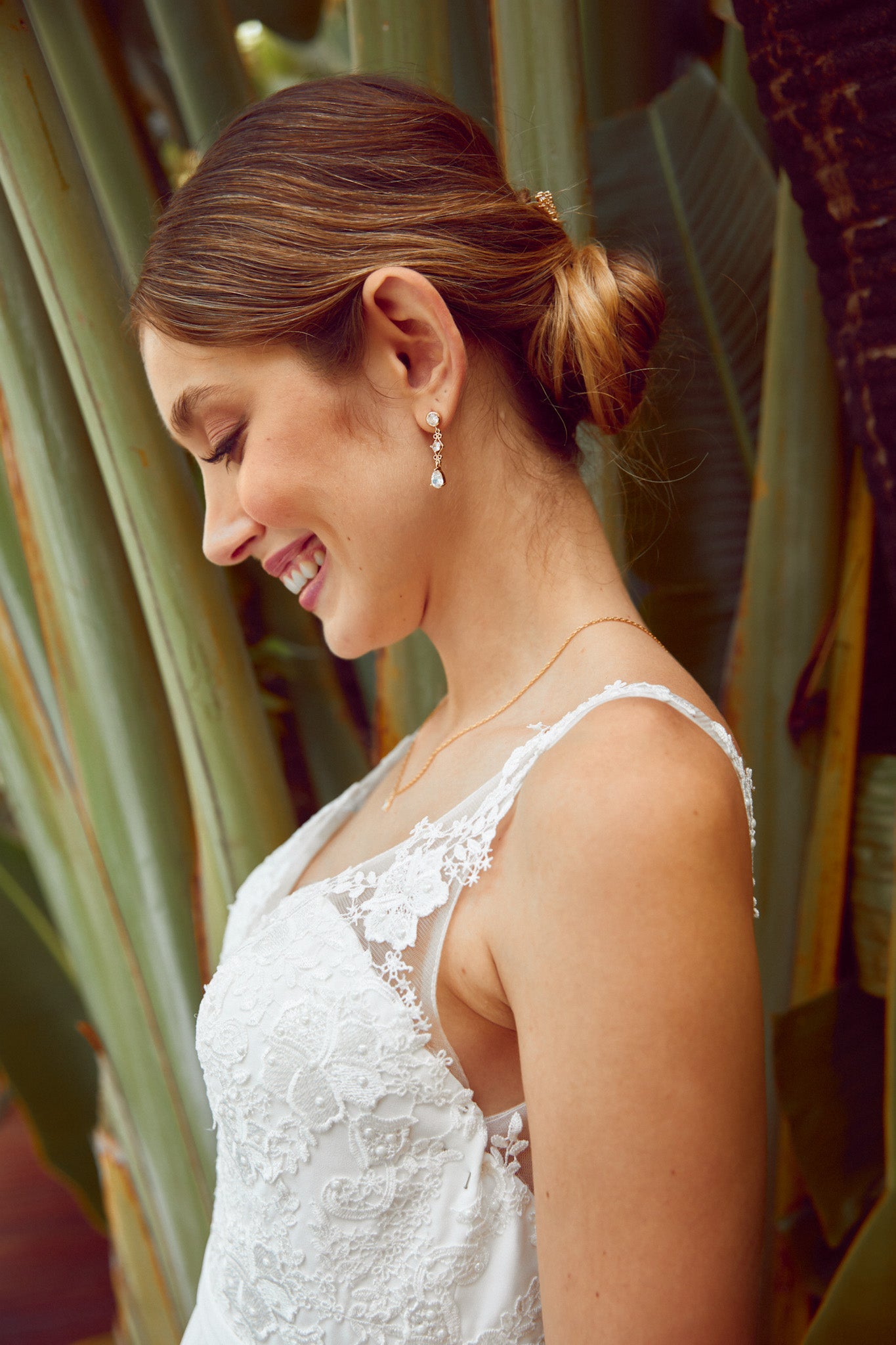 femme de profil souriante qui porte une robe de mariée en dentelle blanche avec des bijoux de mariage romantique en zircons