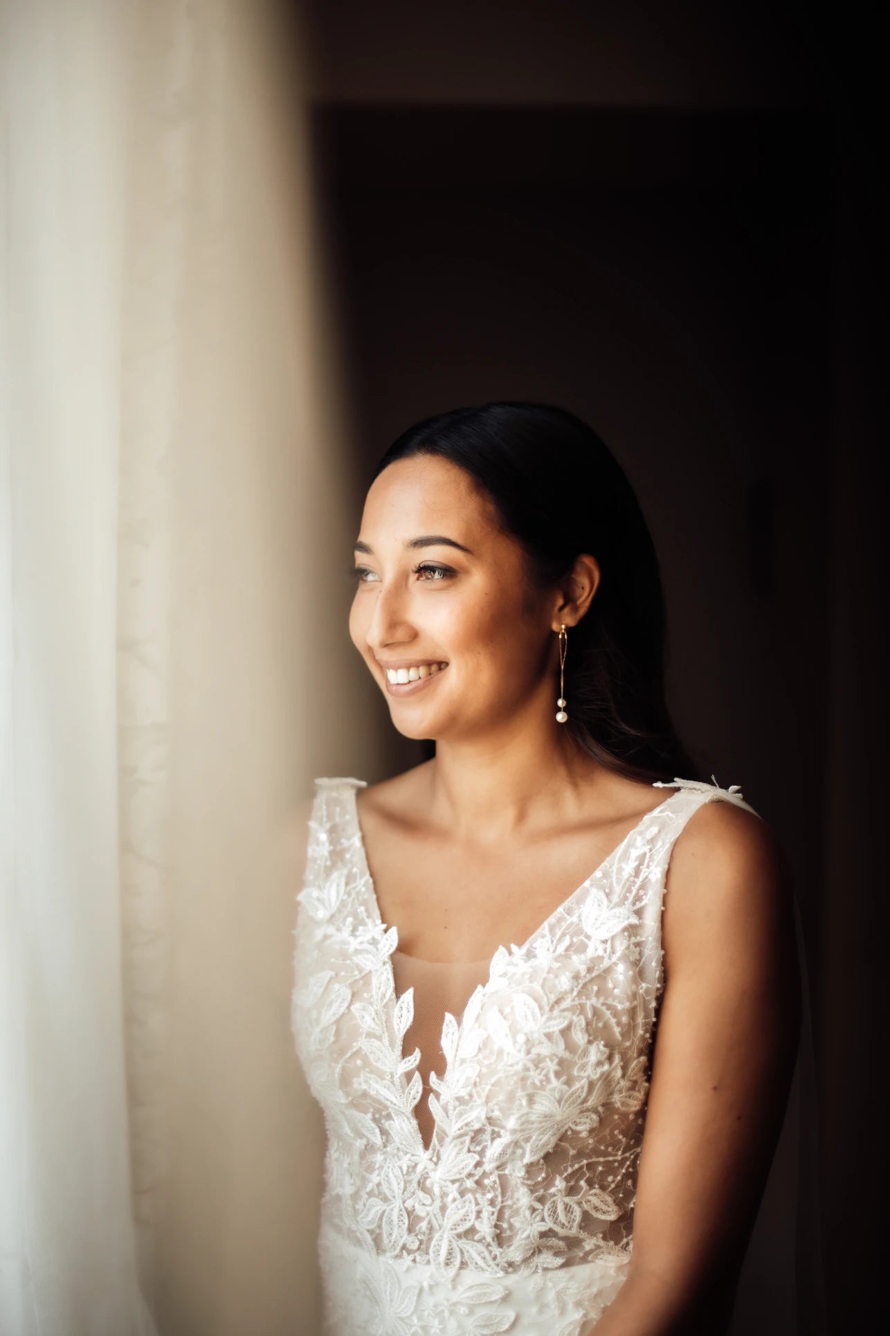 Femme de profil souriant dans sa robe de mariée montrant ses boucles d'oreilles de mariage à perles naturelles