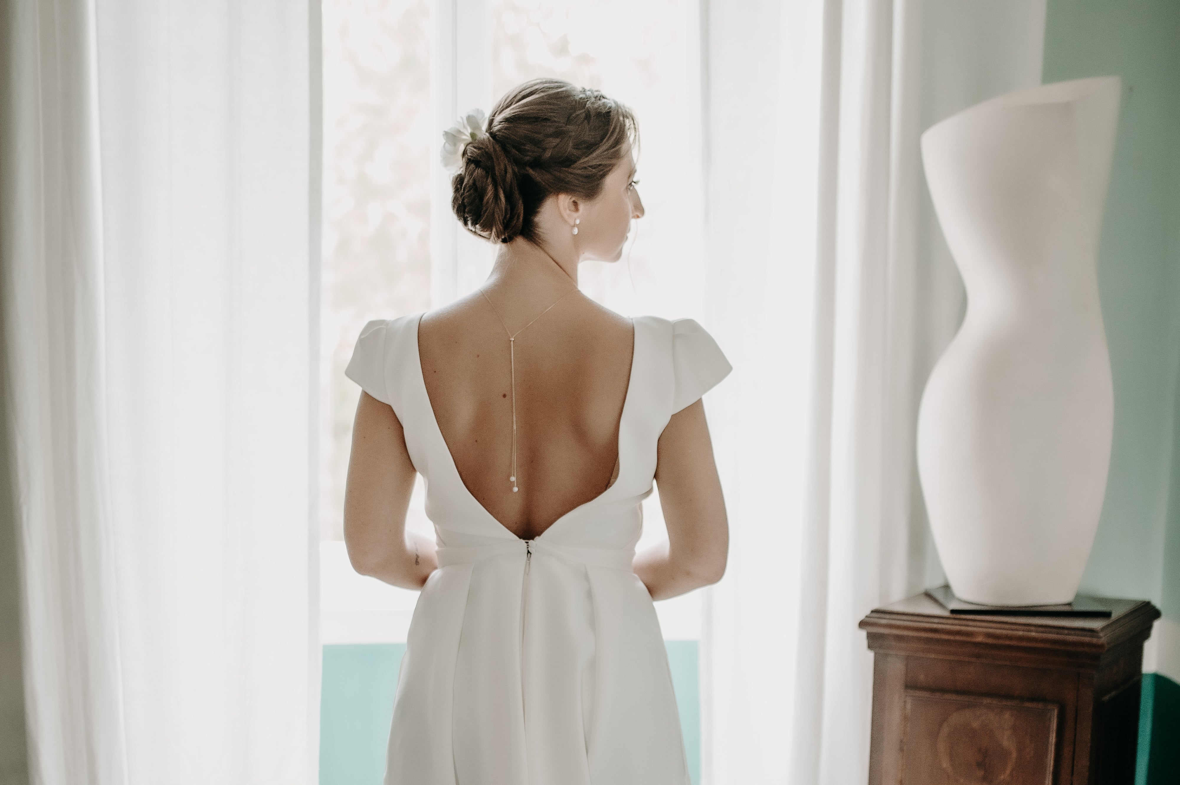 Femme de dos portant une robe de mariage avec un décolleté de dos plongeant et des bijoux pour mariées notamment un collier de dos en perles naturelles