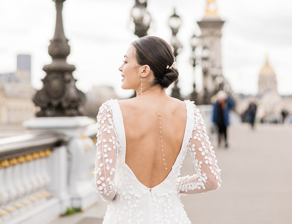 Mariée avec un robe blanche à manches transparentes et décolleté de dos portant des boucles d'oreilles pendantes en perle, un bijoux de tête en perle et un collier de dos en perle sur un pont Parisien