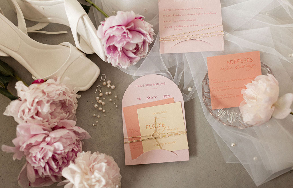 flatlay mariage avec l'invitation du mariage, des pivoines, des chaussures à talons blanches ainsi que des boucles d'oreilles mariage en perles naturelles