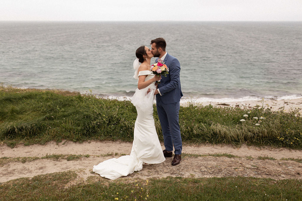 couple devant la plage qui s'embrasse habillé en tenue de mariage avec une robe blanche et un costume bleu marine