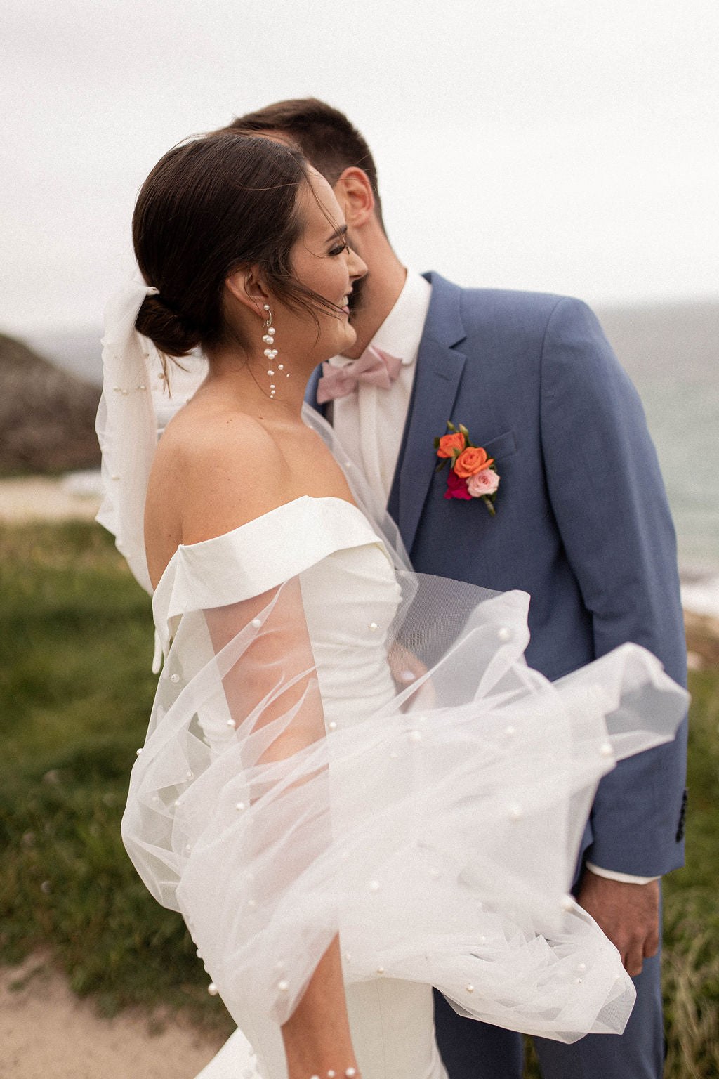 couple le jour du mariage devant la mer, la femme est devant l'homme habillé en robe blanche avec un voile en tuile et perles