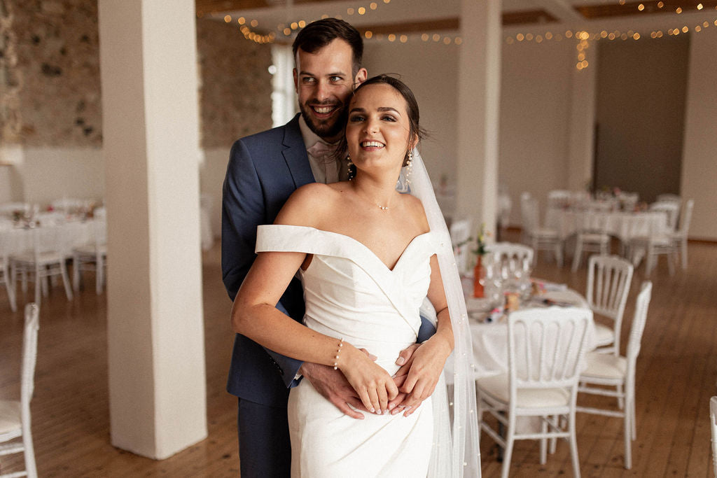 couple qui s'enlace, l'homme est derrière et la femme est devant lui, ils sourient et sont dans la salle de réception du mariage