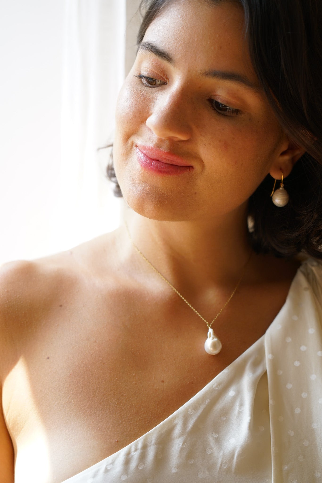 Jeune mariée souriant en regardant en bas montrant sont collier pendant en chaine avec une perle baroque au bout