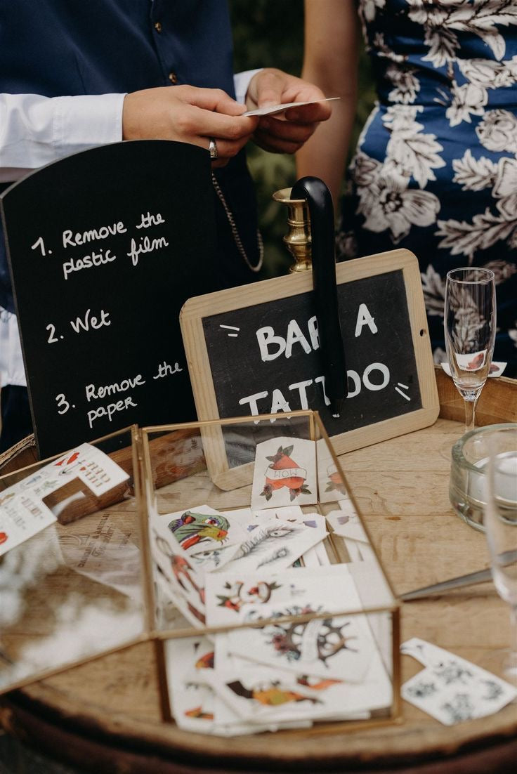 table avec une boite en verre remplis de tatouage éphémère et une ardoise noire avec écrit les instructions pour se faire le tatouage lors d'une animation de mariage