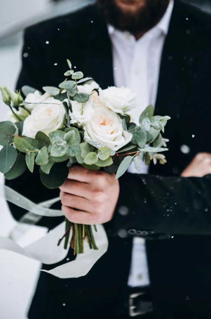 Homme en costume qui va se marier portant à la main le bouquet de fleurs de sa mariée pendant que la neige tombe