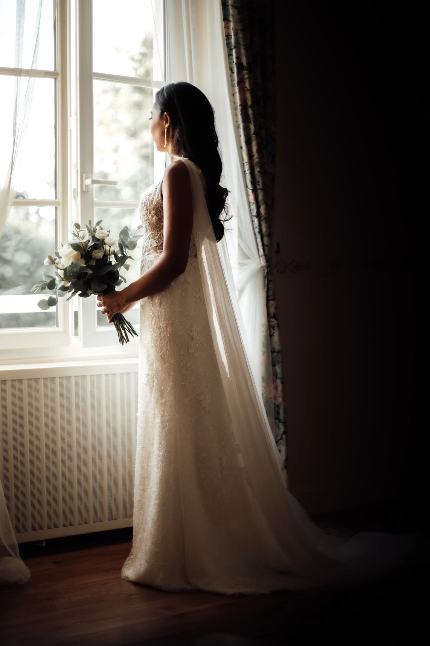 Jeune mariée de profil regardant au loin devant une fenêtre dans sa robe de mariée et ces bijoux de mariage