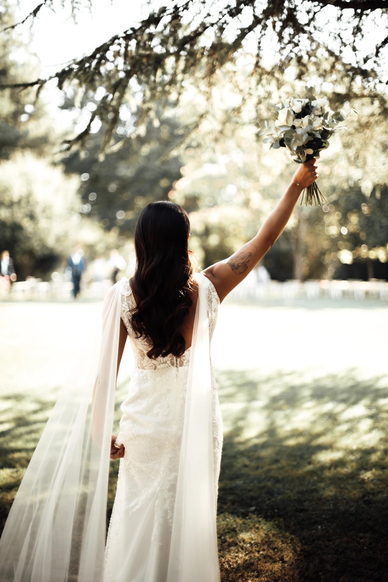 Mariée de dos levant sa main vers le ciel en tenant son bouquet montrant sa robe de mariage à décolleté de dos