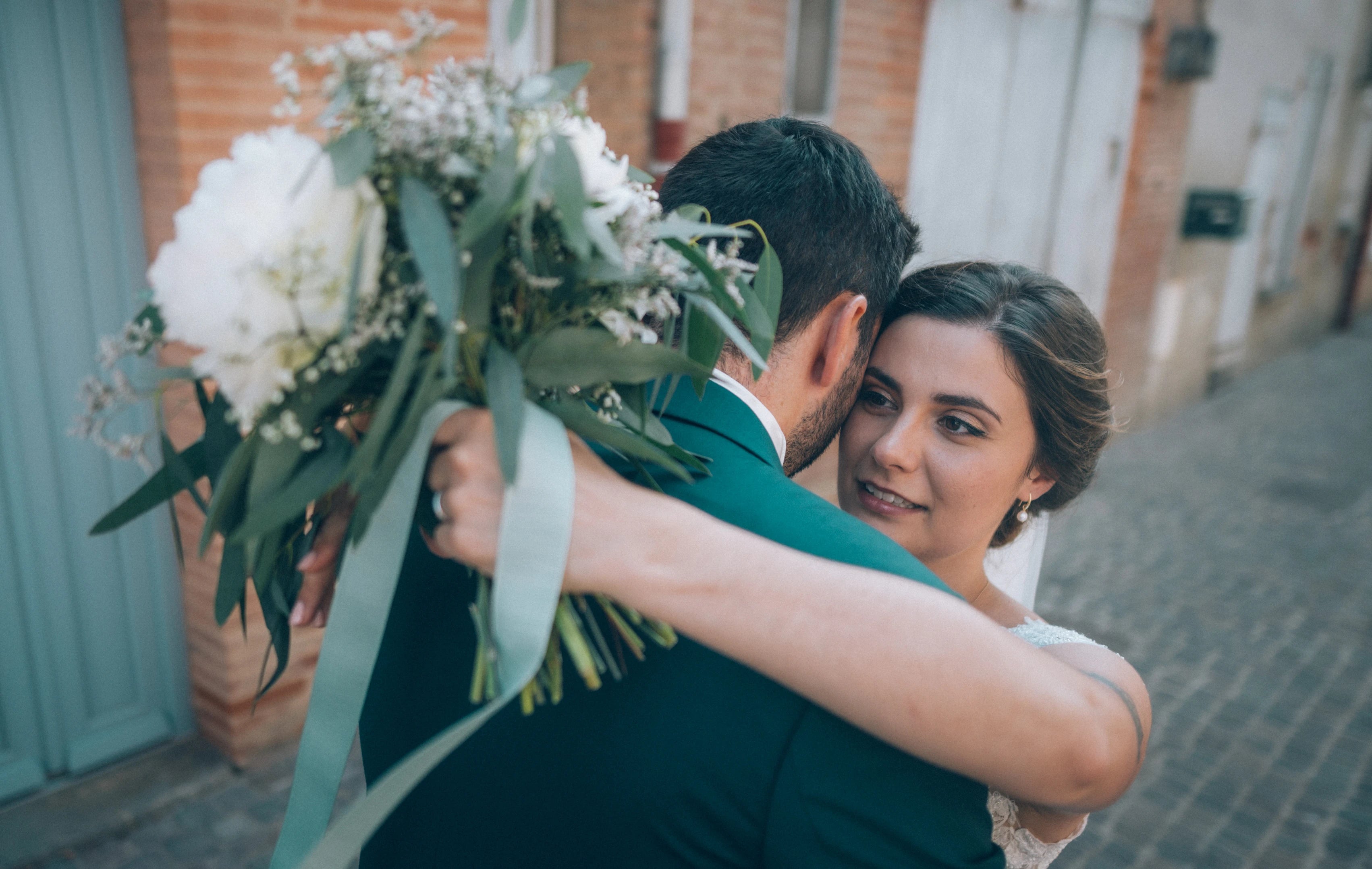 Les mariés qui s'enlacent montrant le visage de la mariée et ses bijoux pour mariées ainsi que son bouquet derrière le dos de son mari 