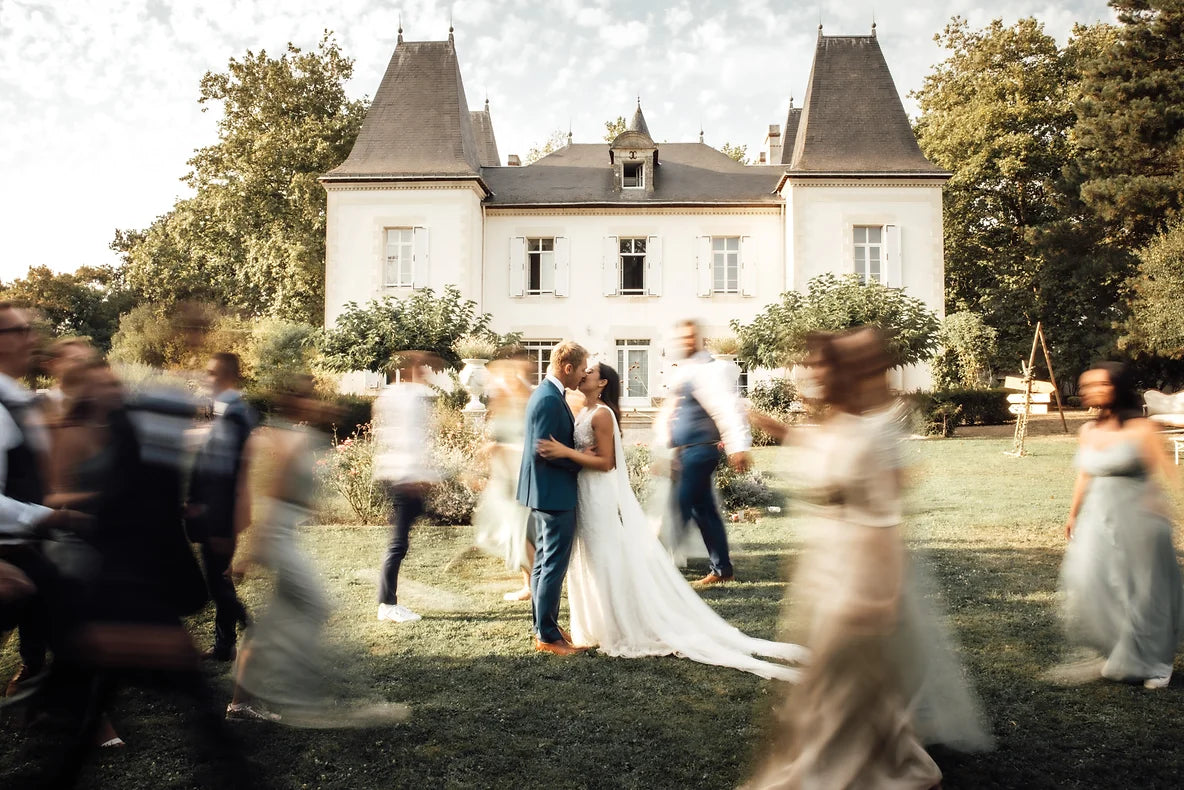 Au centre de la photo deux mariés qui s'embrassent avec les invités qui bougent autour avec un effet de flou