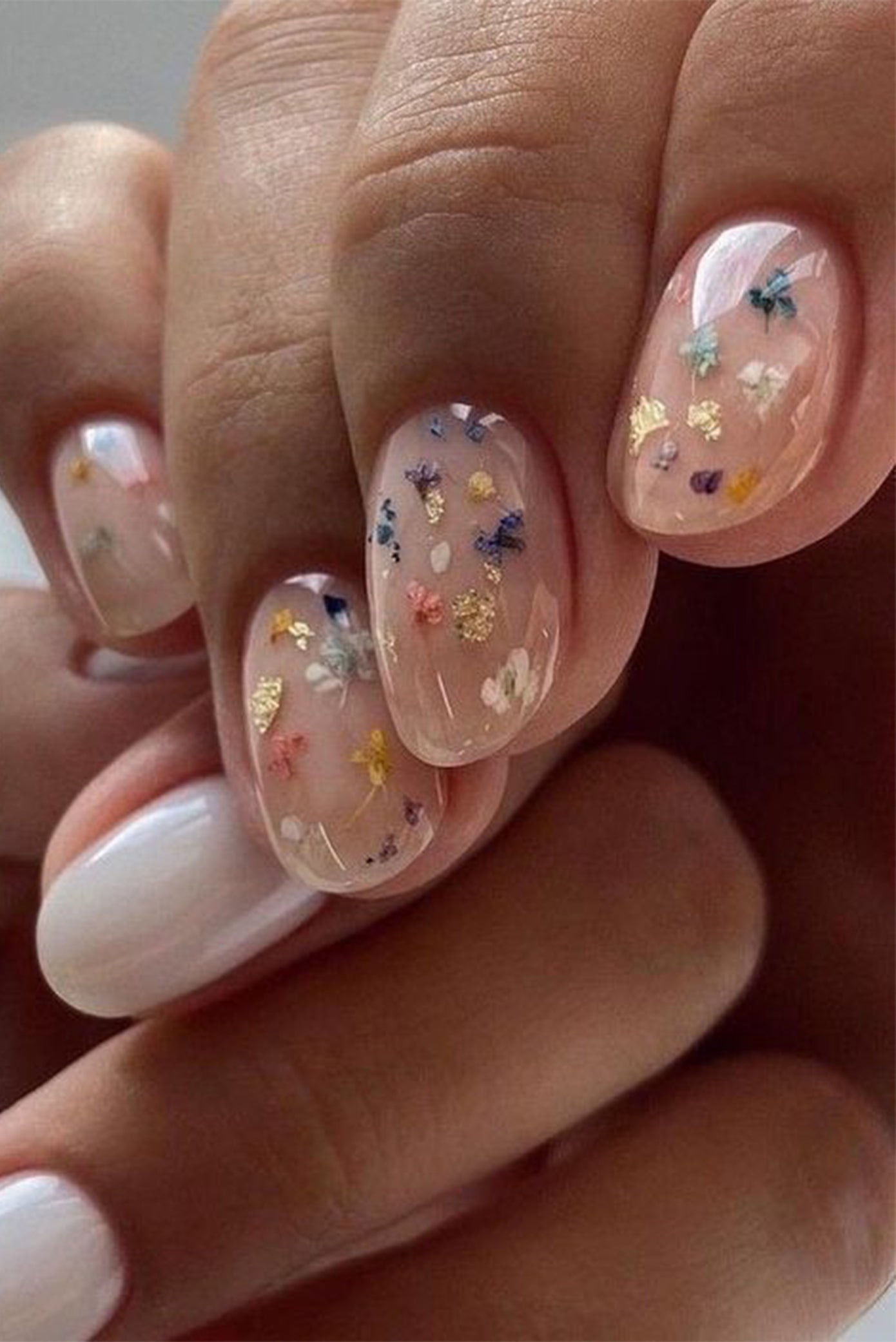 Gros plan sur des ongles manucurés nude avec à l'intérieur des fleurs séchées incrustées de différente couleurs