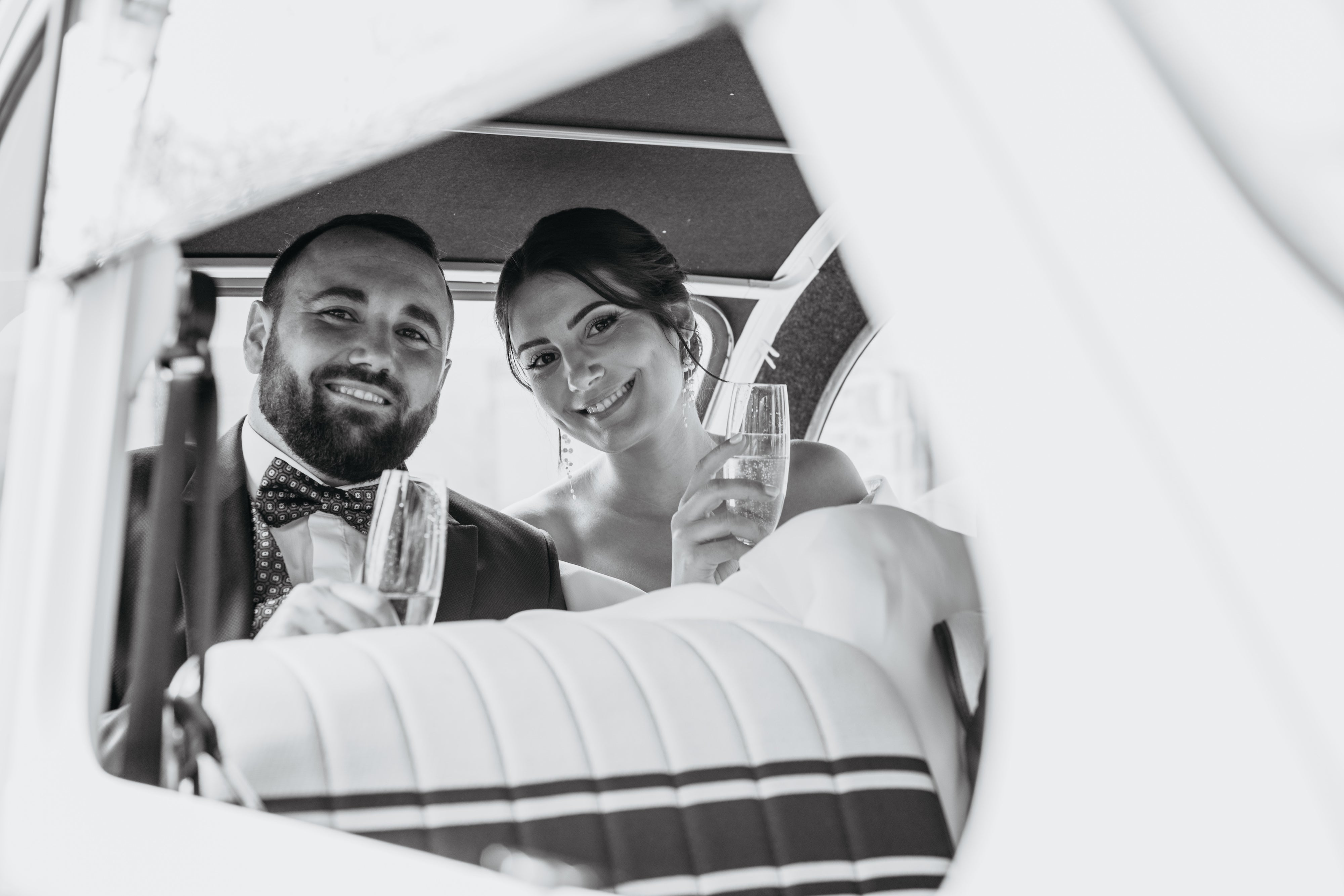 Deux jeune mariés dans une voiture souriant avec une coupe de champagne à la main montrant des bijoux de mariage