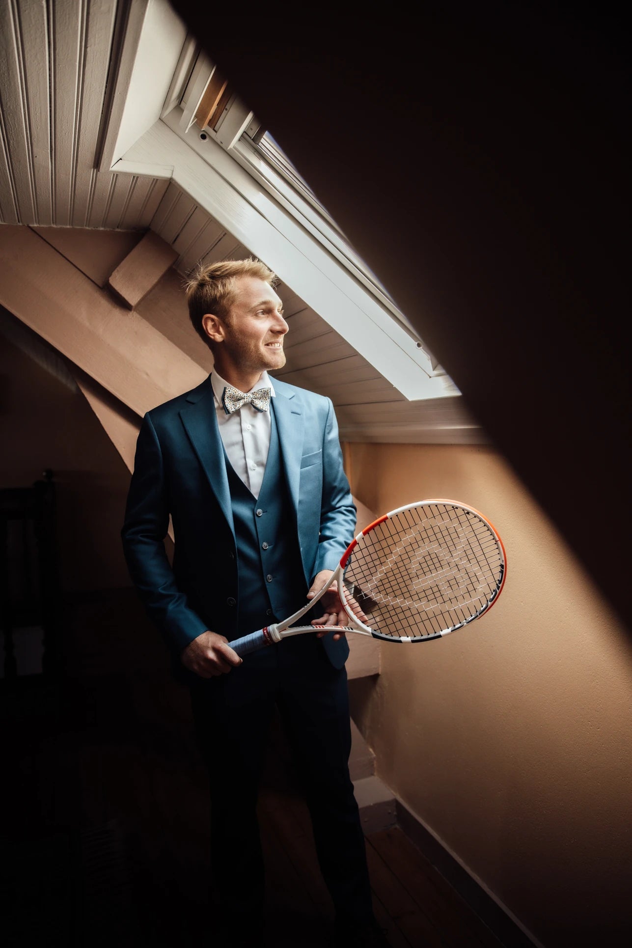 Jeune marié en costume de mariage avec un noeud papillon qui regarde sur le côté en portant une raquette de tennis