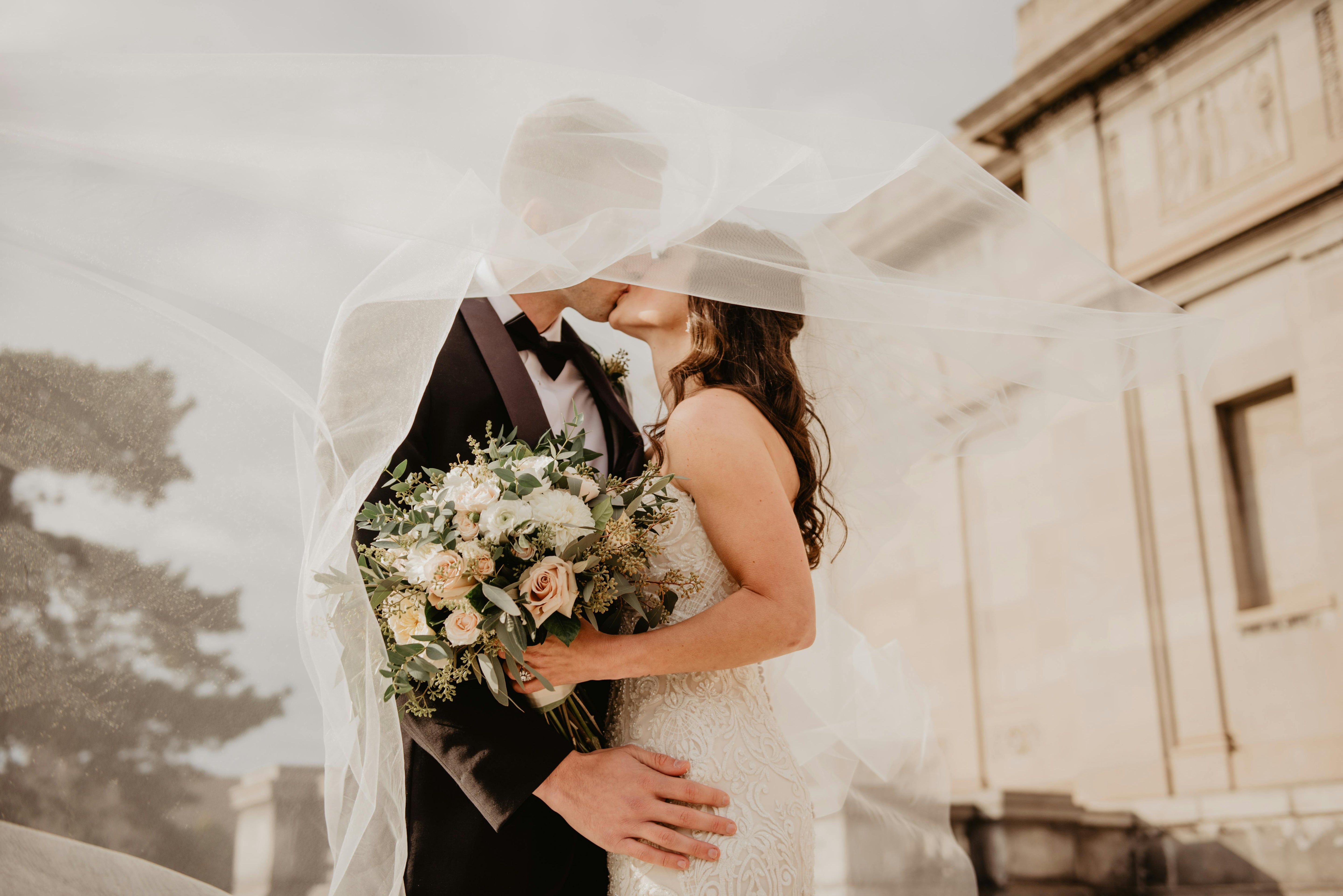 Couple de mariés qui s'embrasse sous le voile de la mariée en portant un bouquet de fleurs à la main