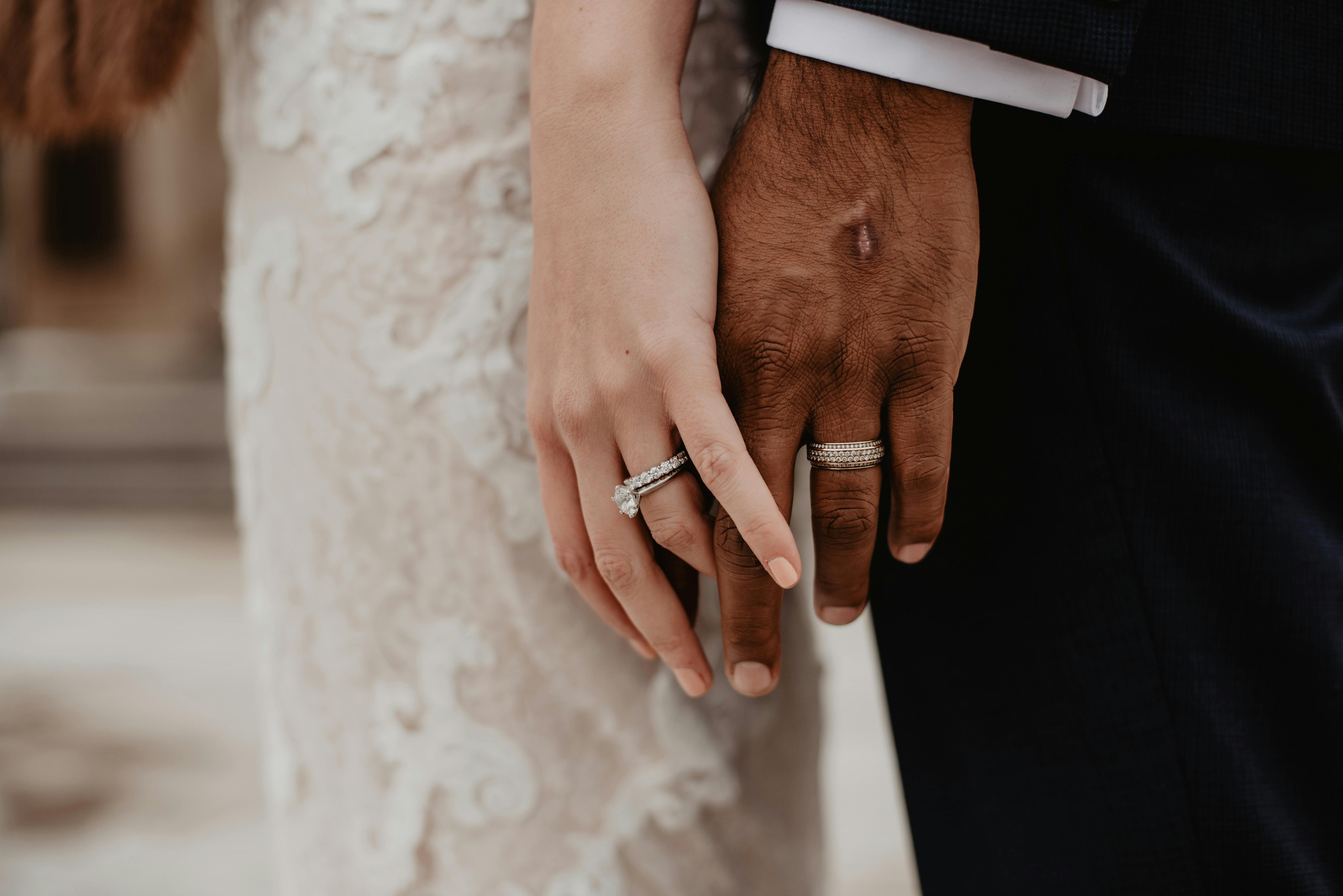 Focus sur la main de deux mariés portant leurs bagues avec en fond leurs tenues de mariage