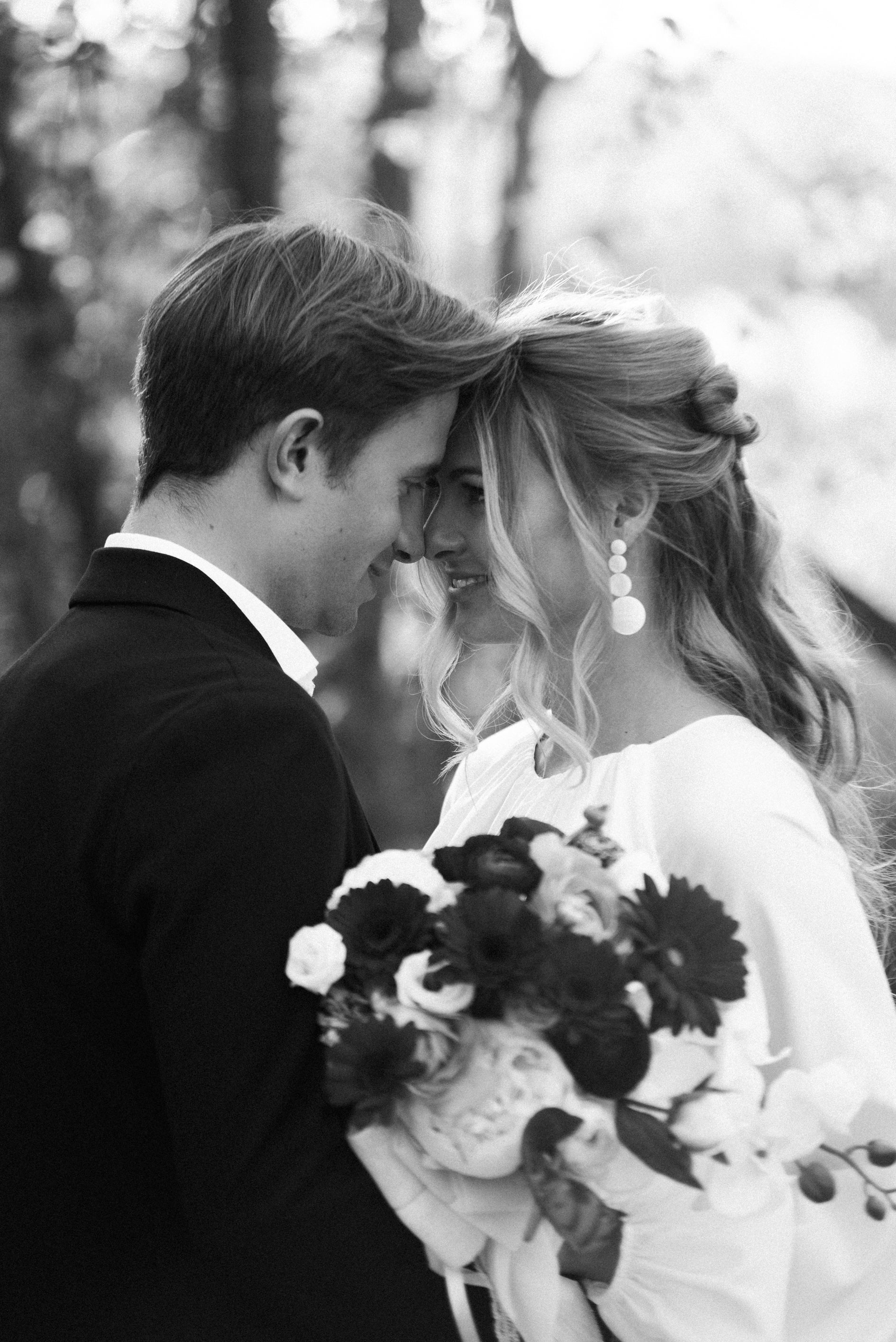 Couple de mariés se regardant front contre front en noir et blanc, mariée portant des boucles d'oreilles pendantes au style bohème et élégant